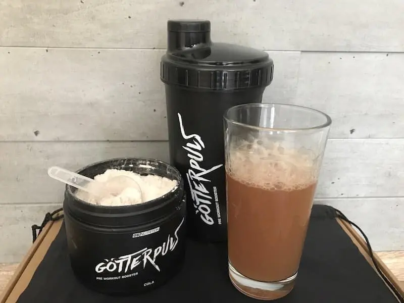 Geöffnete Götterpuls-Dose mit Pulver, Messlöffel, Shaker und angerührtem Cola-Drink